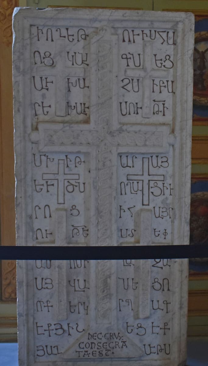 White marble khachkar carved with a large cross and, in the corners, blocky Armenian capital letters incised into the stone