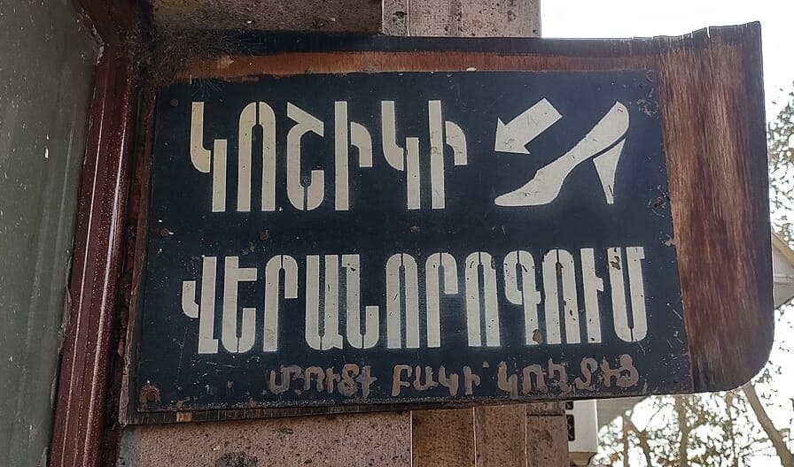 Weathered black metal sign with hand-painted white Armenian capital letters and a small arrow and shoe icon, mounted on a stone pillar