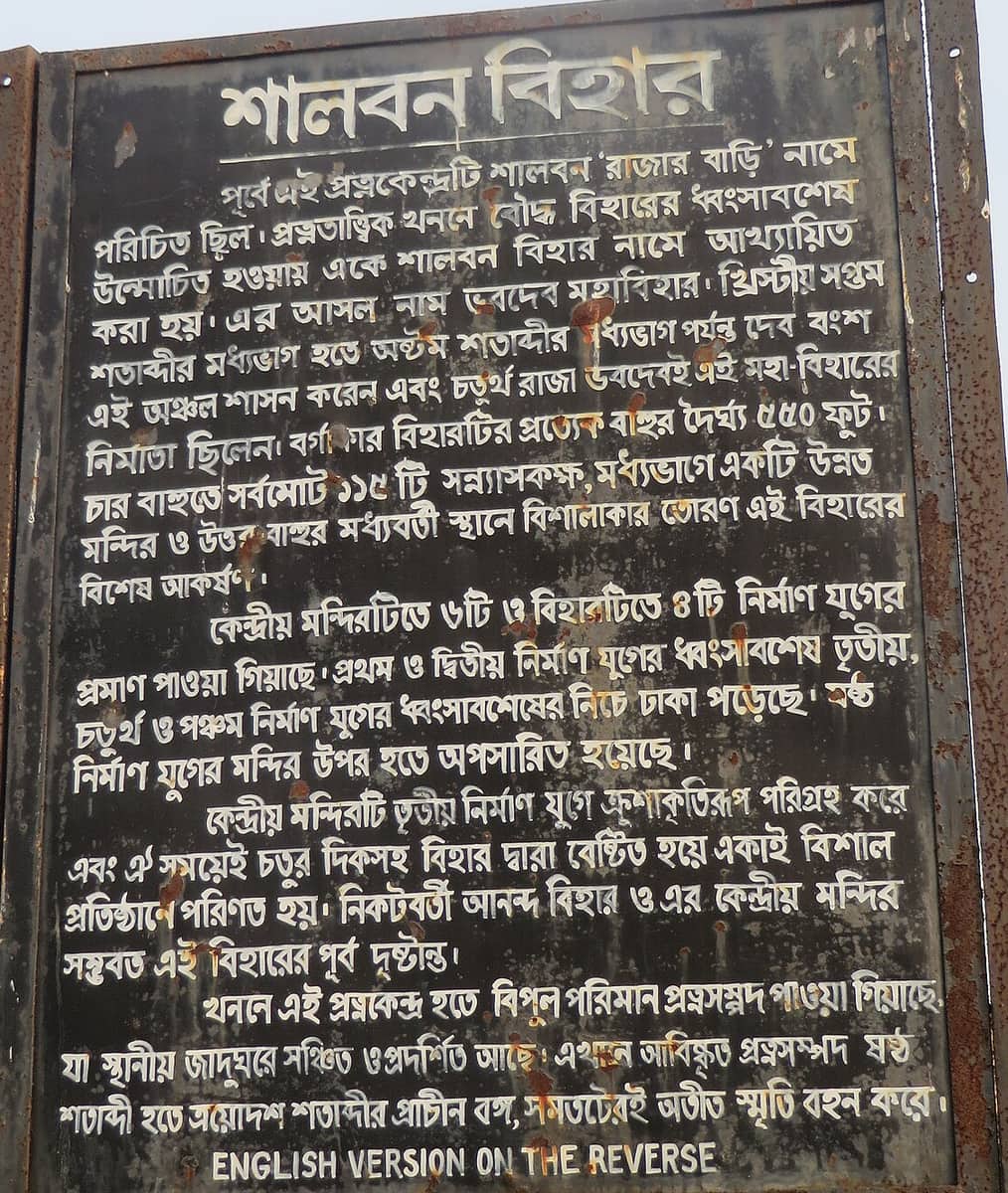 Weathered dark metal information board covered in painted Bengali text, photographed close up against a pale sky