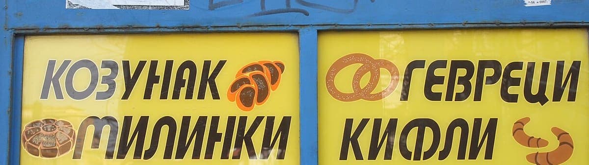 Blue kiosk window with two yellow signs in bold black capitals advertising kozunak, milinki, gevreci and kifli, each beside a small illustration of the pastry