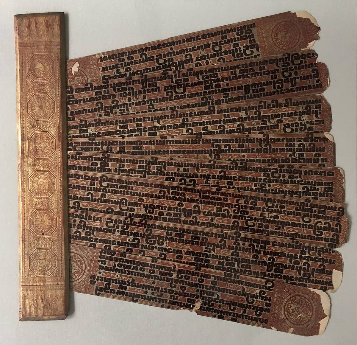 A fanned stack of gilded palm-leaf manuscript pages bordered in red lacquer with gold roundels, beside a matching gilded wooden cover