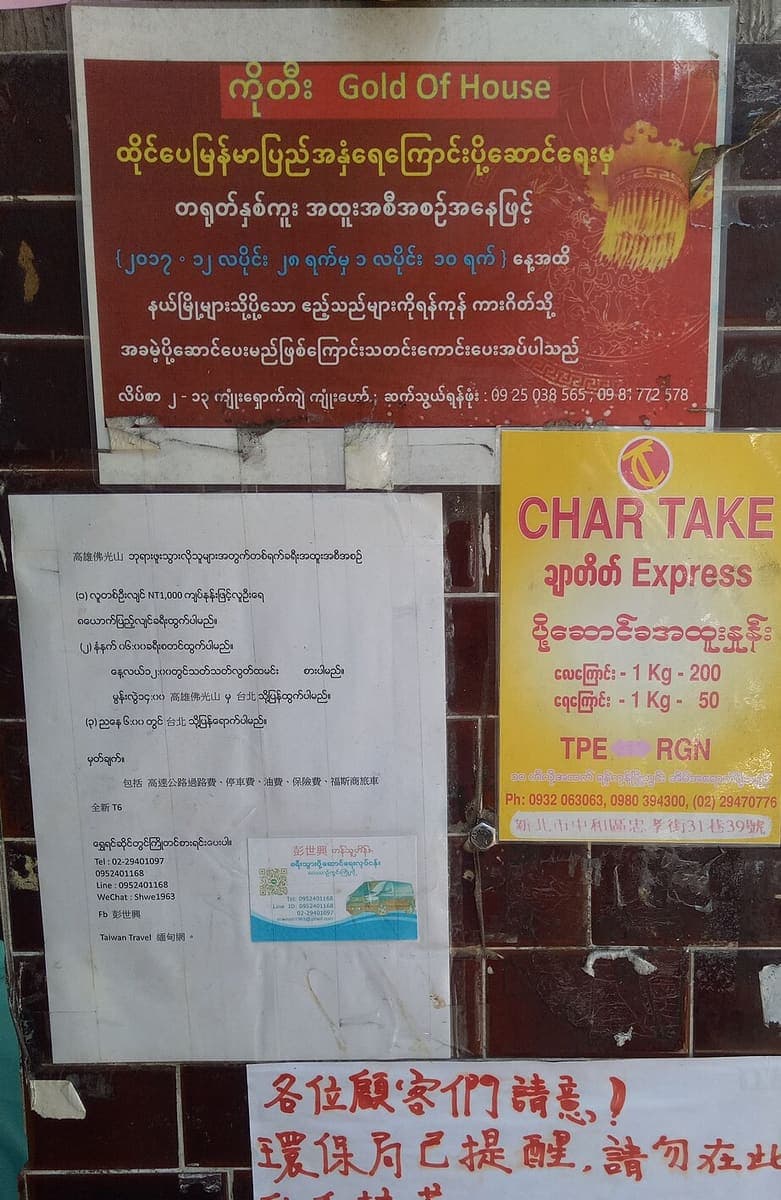 A tiled wall covered with printed and handwritten notices in Burmese and Chinese, including a red gold-shop advertisement and a yellow courier-service poster