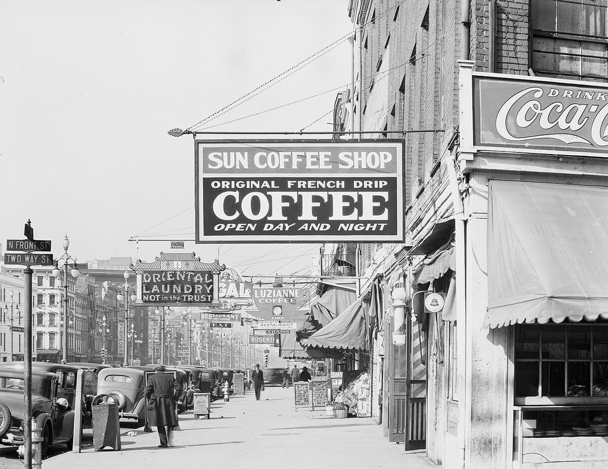 A 1930s street photograph with a large hanging shop sign in the foreground and many smaller painted signs receding down the block