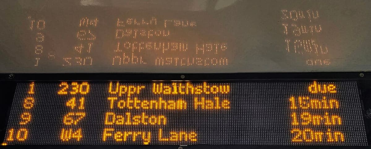 An orange dot-matrix bus countdown display showing four rows of stand number, route, destination and waiting time, mirrored in the shelter roof above
