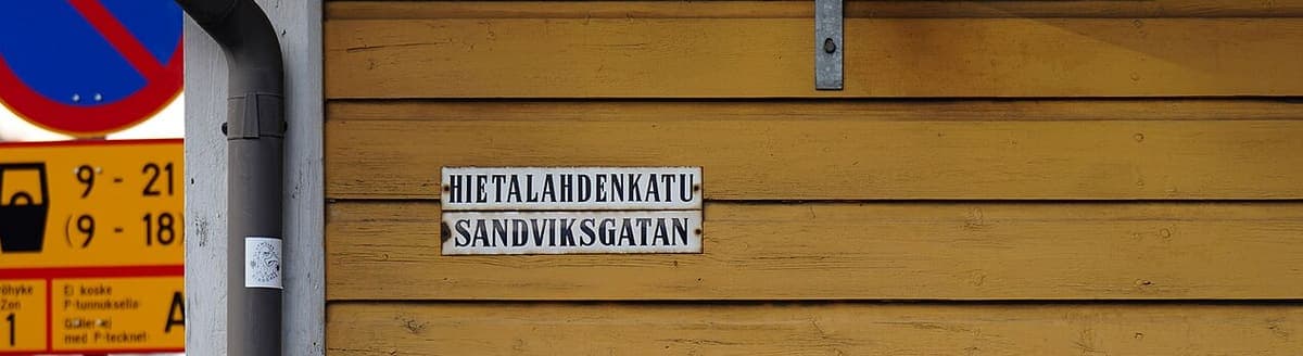Bilingual metal street sign reading Hietalahdenkatu above Sandviksgatan on a yellow wooden building in Helsinki