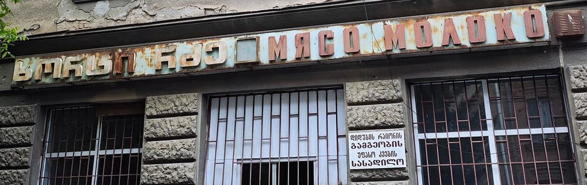 Rusted metal shop fascia in Tbilisi with raised Georgian and Russian lettering, and a small white plaque with four lines of Georgian below