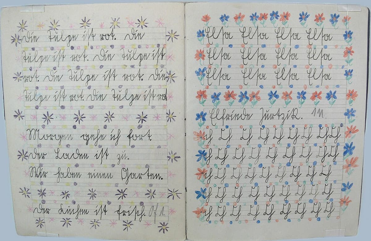 An open school exercise book showing two ruled pages of German cursive practice, each framed by a border of hand-drawn coloured-pencil flowers