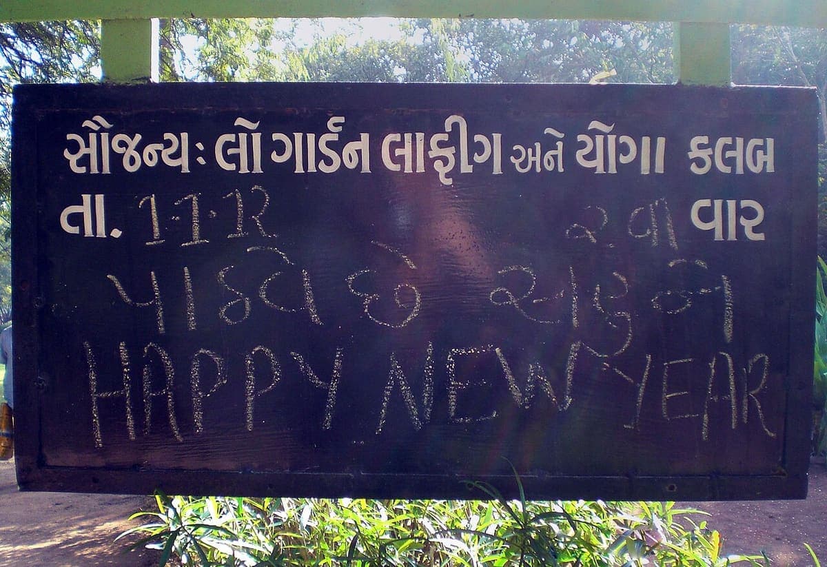A dark wooden signboard with Gujarati letters painted in white, a date in Gujarati-style numerals chalked into the blanks, and "HAPPY NEW YEAR" chalked underneath in Latin letters