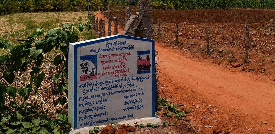 A small blue and white concrete signboard giving details of a rural roadworks scheme in Kannada, standing beside a red-earth farm track between a fenced plantation and a ploughed field