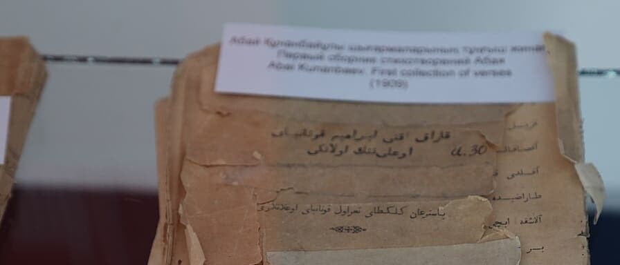 A torn, taped fragment of an old Arabic-script Kazakh book title page, photographed under museum glass, with more Arabic-script lines showing on the leaf beside it