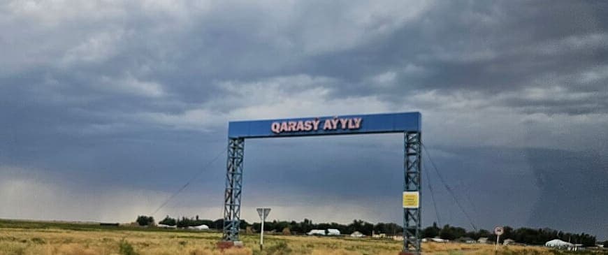 A blue steel gantry on lattice towers at the edge of a village, with QARASÝ AÝYLY spelled out in Latin capitals under a stormy sky