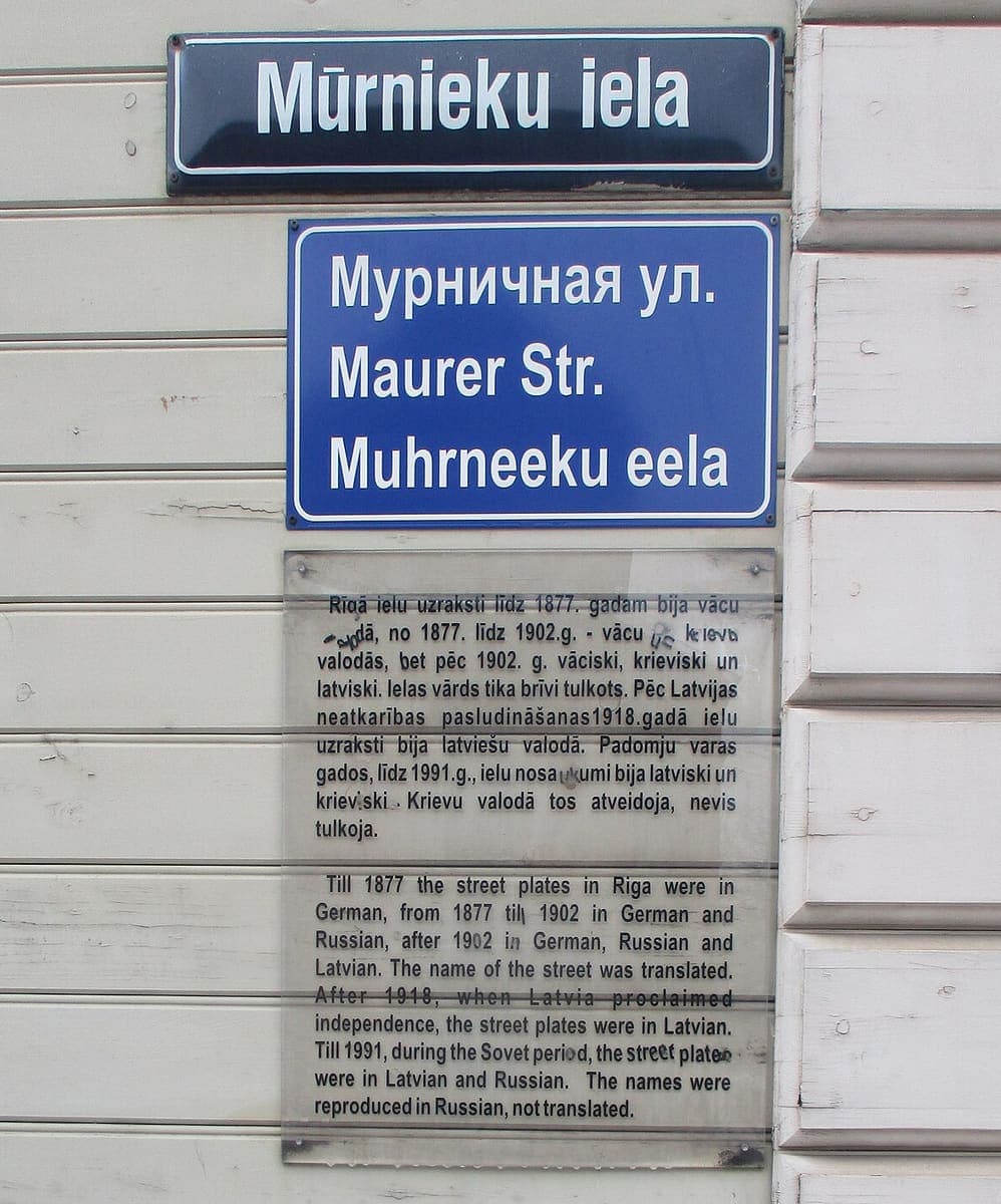 Three signs stacked on a wall: a modern blue Latvian street sign reading Mūrnieku iela, an older-style plate giving the street name in Russian, German and old-orthography Latvian, and an explanatory plaque in Latvian and English
