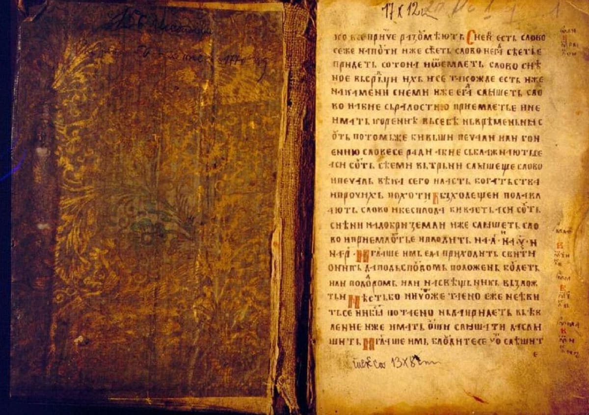 Open manuscript with an ornately patterned leather cover on the left page and a page of red-and-black Cyrillic text with marginal notes on the right
