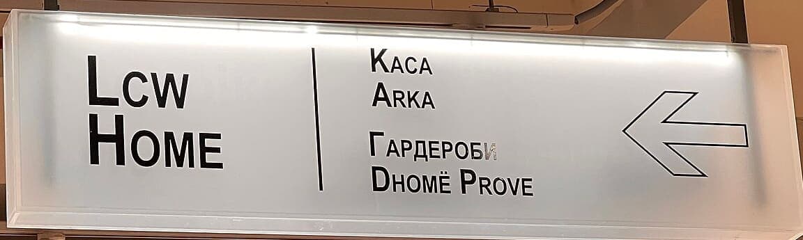 Illuminated mall directional sign with an arrow, giving a shop name and section labels in English, Macedonian Cyrillic and Albanian