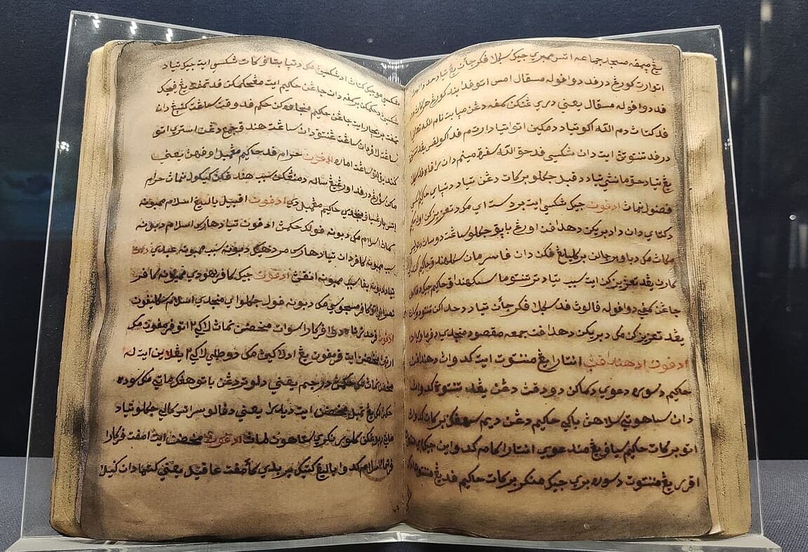 An open manuscript book with dense Jawi script in black ink and red rubricated section markers, resting on a clear museum stand