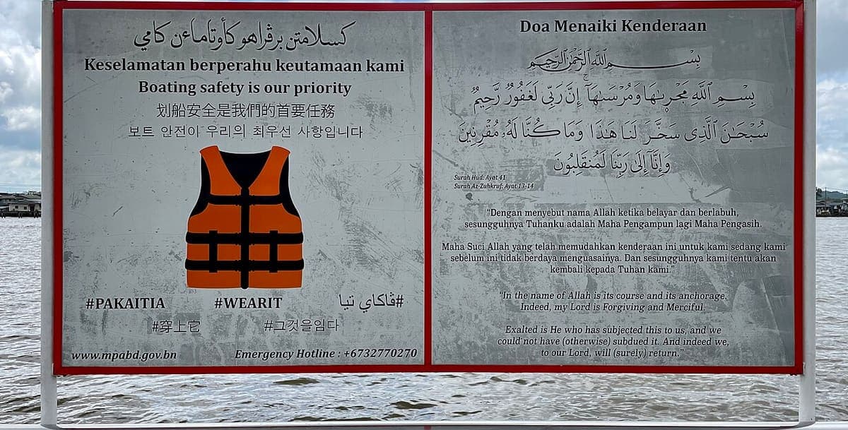 A weathered metal noticeboard beside a waterway, printed with a life-jacket safety notice in Jawi, Rumi Malay, English, Chinese and Korean