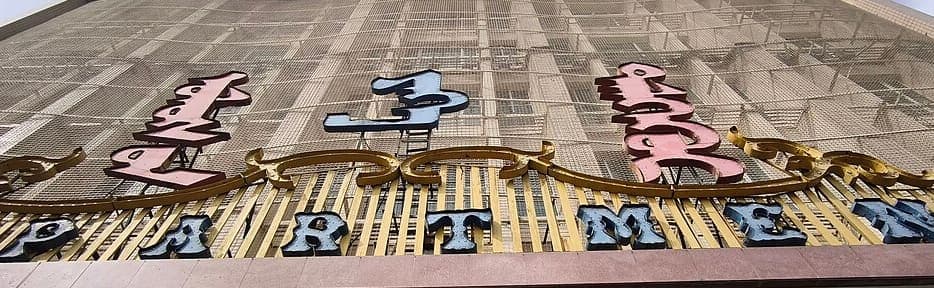 Three large ornamental Mongolian script words in pink and pale blue, mounted on a gilded scroll high on a building facade above Latin letters reading PARTMEN