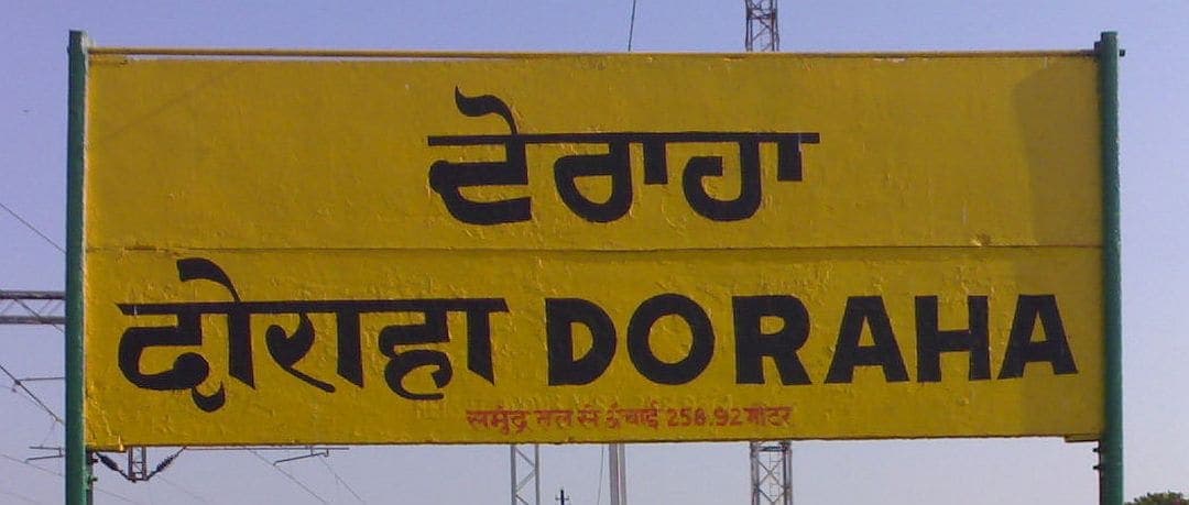 A yellow railway station nameboard giving the same place name in Gurmukhi on the upper line and in Devanagari and Latin letters on the line below