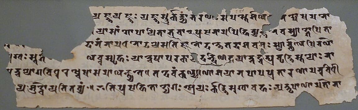 Torn strip of pale manuscript with six lines of angular Sanskrit writing, large pieces missing from the left and upper edges