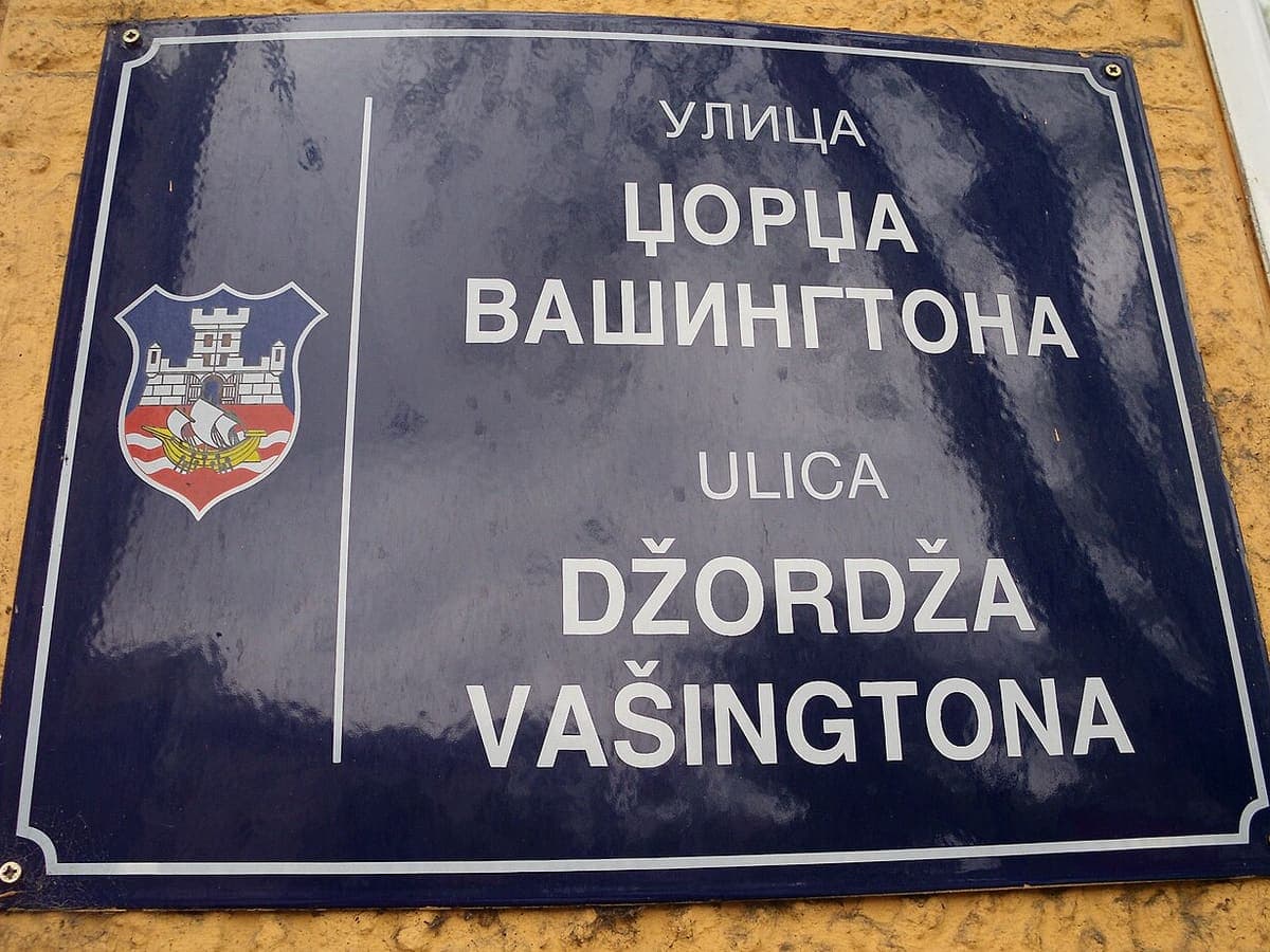 Dark blue enamel street sign with the city arms on the left and the same street name given twice, first in Cyrillic then in Latin script