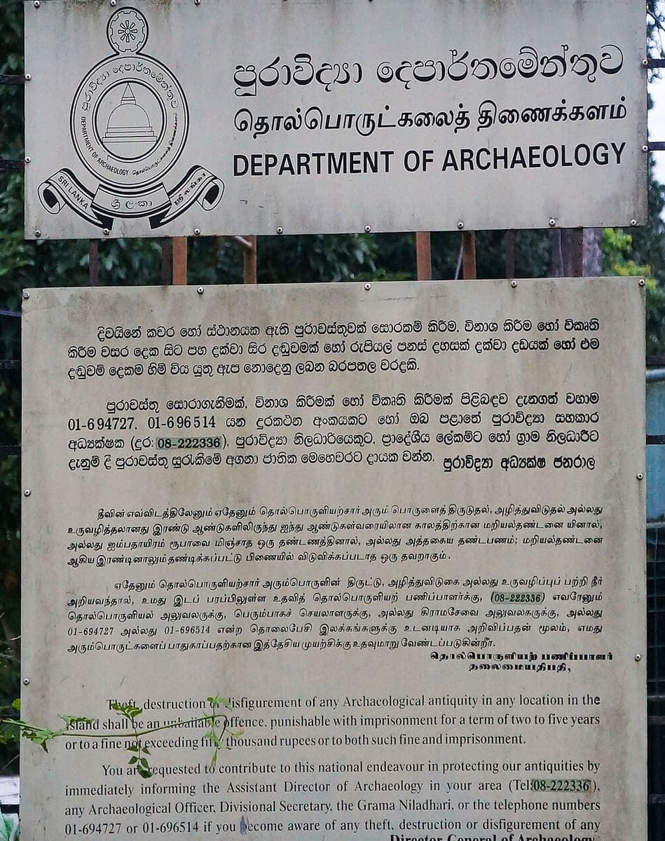 Metal heritage-protection notice with two printed paragraphs of Sinhala text above matching Tamil and English versions, mounted below a Department of Archaeology sign