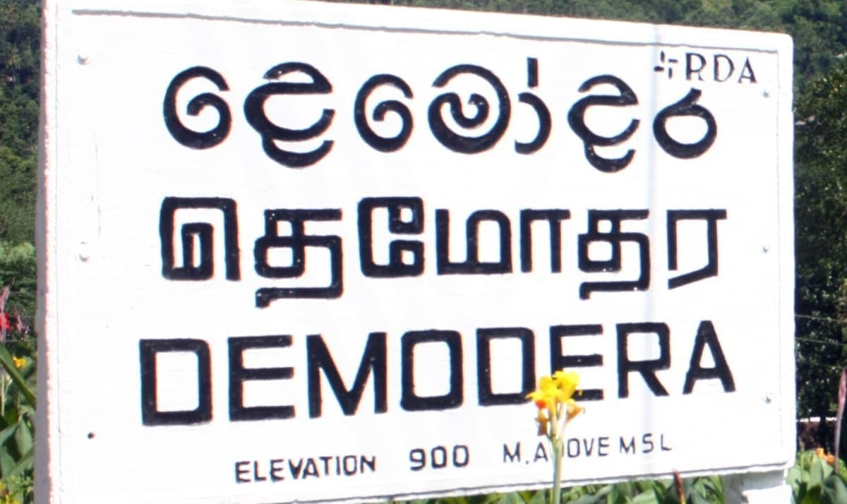 White roadside name board reading the same place name in rounded Sinhala script, Tamil script and Latin letters, with an elevation line underneath
