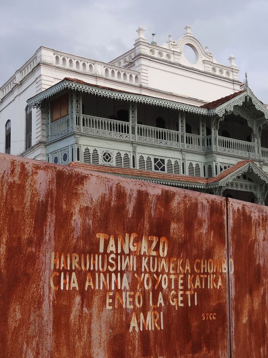 A Swahili notice headed TANGAZO, hand-painted in pale letters on a rusted metal fence in front of an ornate carved Zanzibari balcony