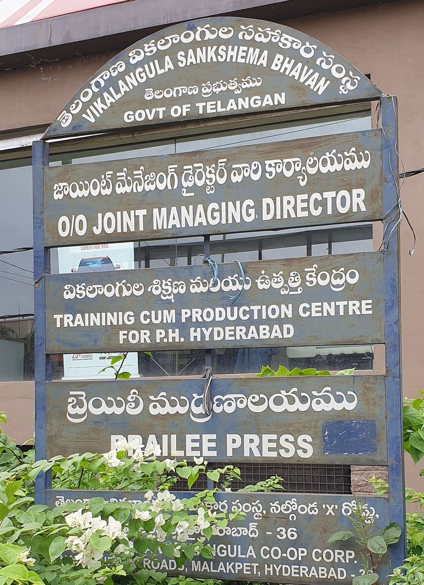 Weathered multi-panel government signboard in Telugu and English naming a welfare cooperative, its managing director’s office, a training centre and a Braille press