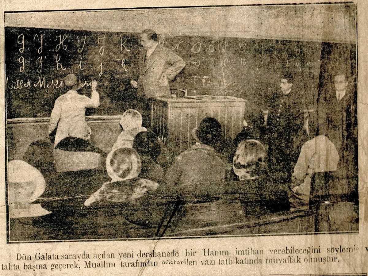 Grainy newspaper photograph of a classroom with new Latin letters chalked on a blackboard and a teacher standing beside a pupil at the board, with a caption printed below