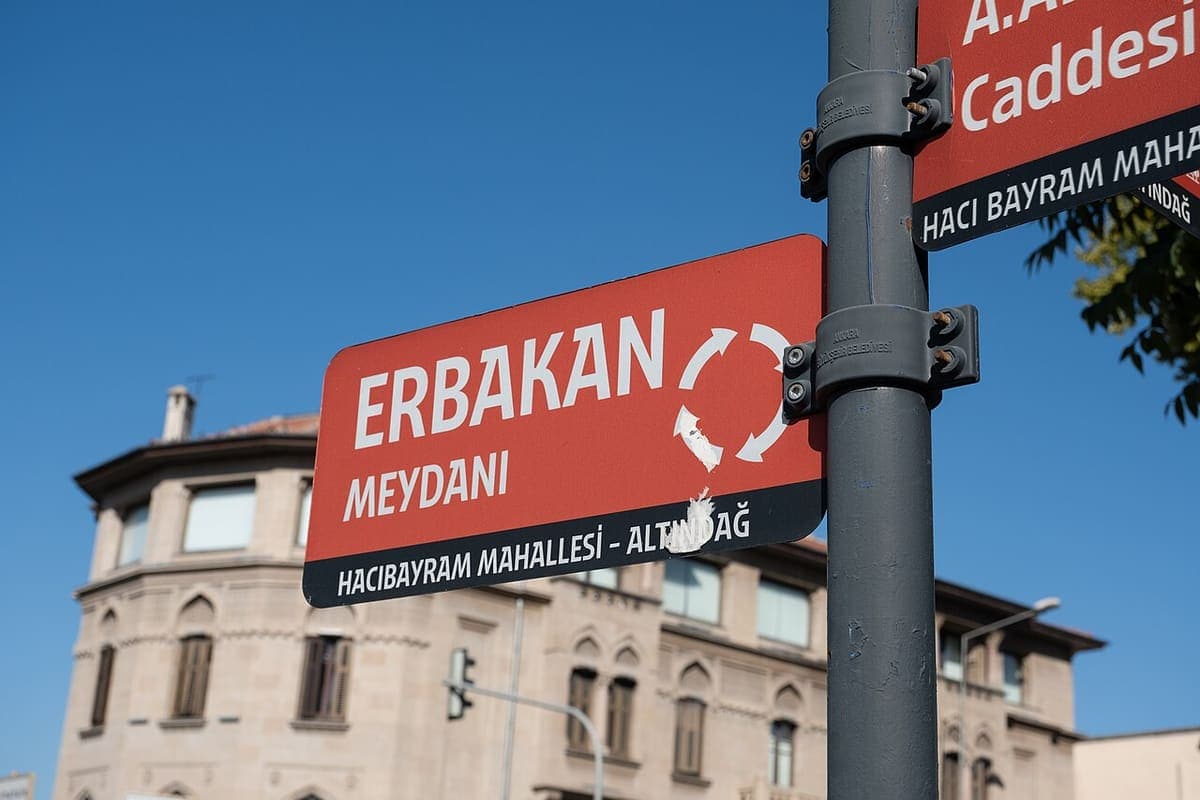 Red municipal street-name sign reading Erbakan Meydanı, Hacıbayram Mahallesi - Altındağ, mounted on a pole beneath a second street sign