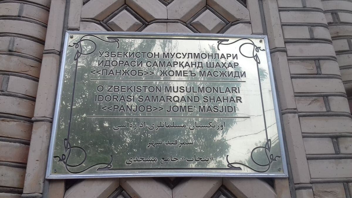 A polished metal plaque naming a mosque in Samarkand, its text set out in three stacked blocks: Cyrillic, Latin and Perso-Arabic script