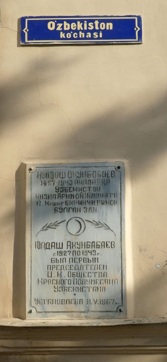 A blue Latin-script street sign reading O'zbekiston ko'chasi above an older engraved memorial plaque in Cyrillic Uzbek and Russian on the same building wall