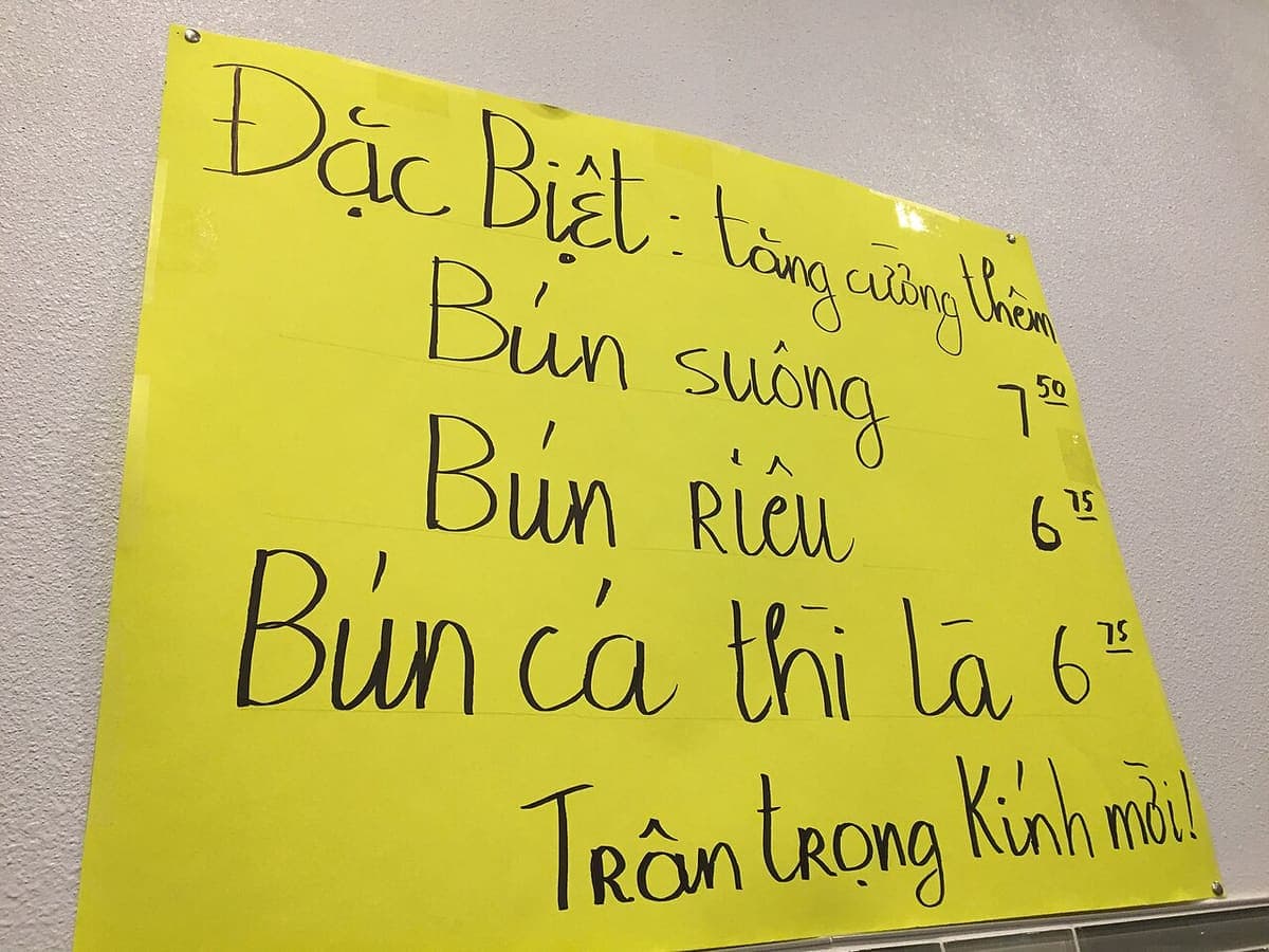 Handwritten yellow sign in Vietnamese listing three noodle dishes with prices, photographed at an angle inside a restaurant