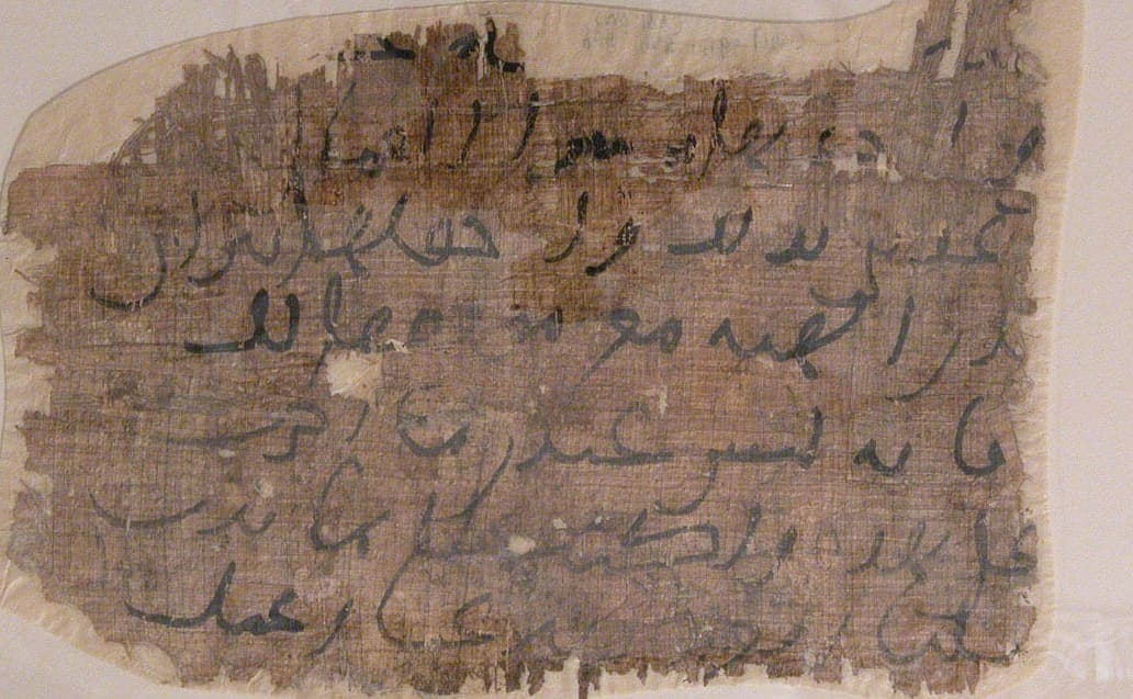 Close view of a torn papyrus fragment with six widely spaced lines of thick black Arabic writing and no consonantal dots