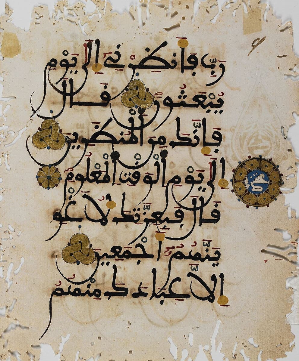 Worm-eaten parchment leaf with seven lines of heavy black Maghribi script, gold trefoil verse markers and a blue and gold marginal medallion