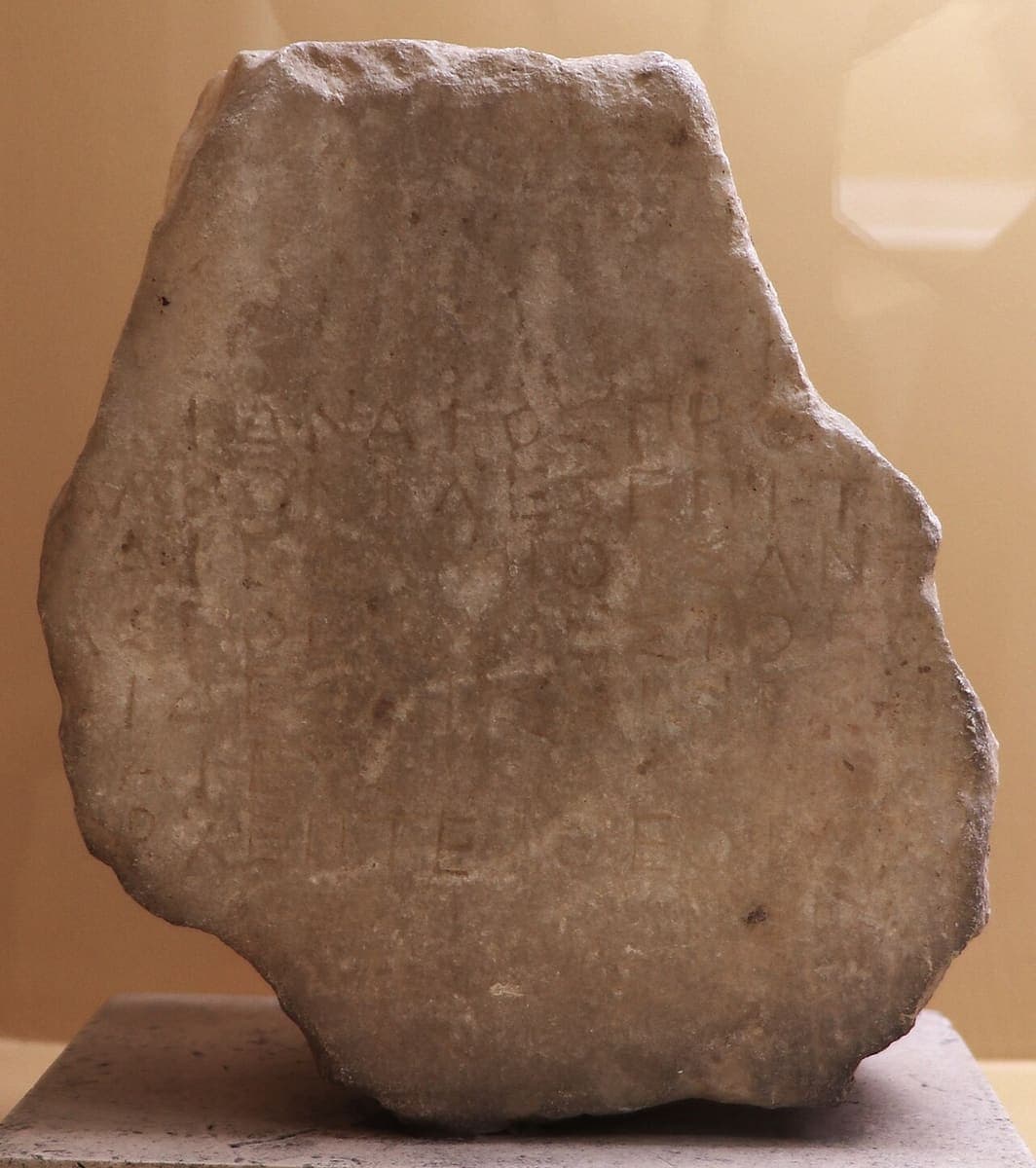 Irregular marble fragment with rows of small worn Greek capitals in a stoichedon grid, photographed against a beige backdrop