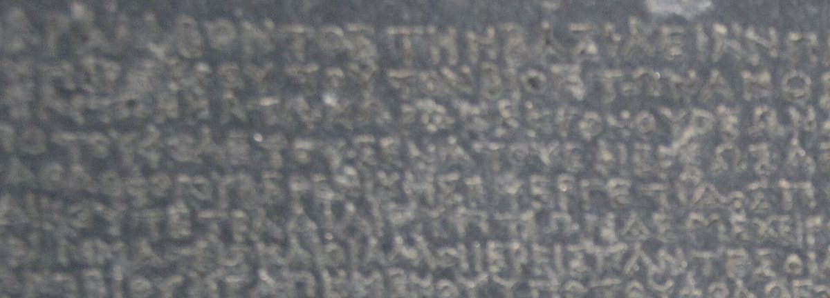Dense rows of small evenly cut Greek capitals on dark granodiorite, the beginning of the Greek section of the Rosetta Stone
