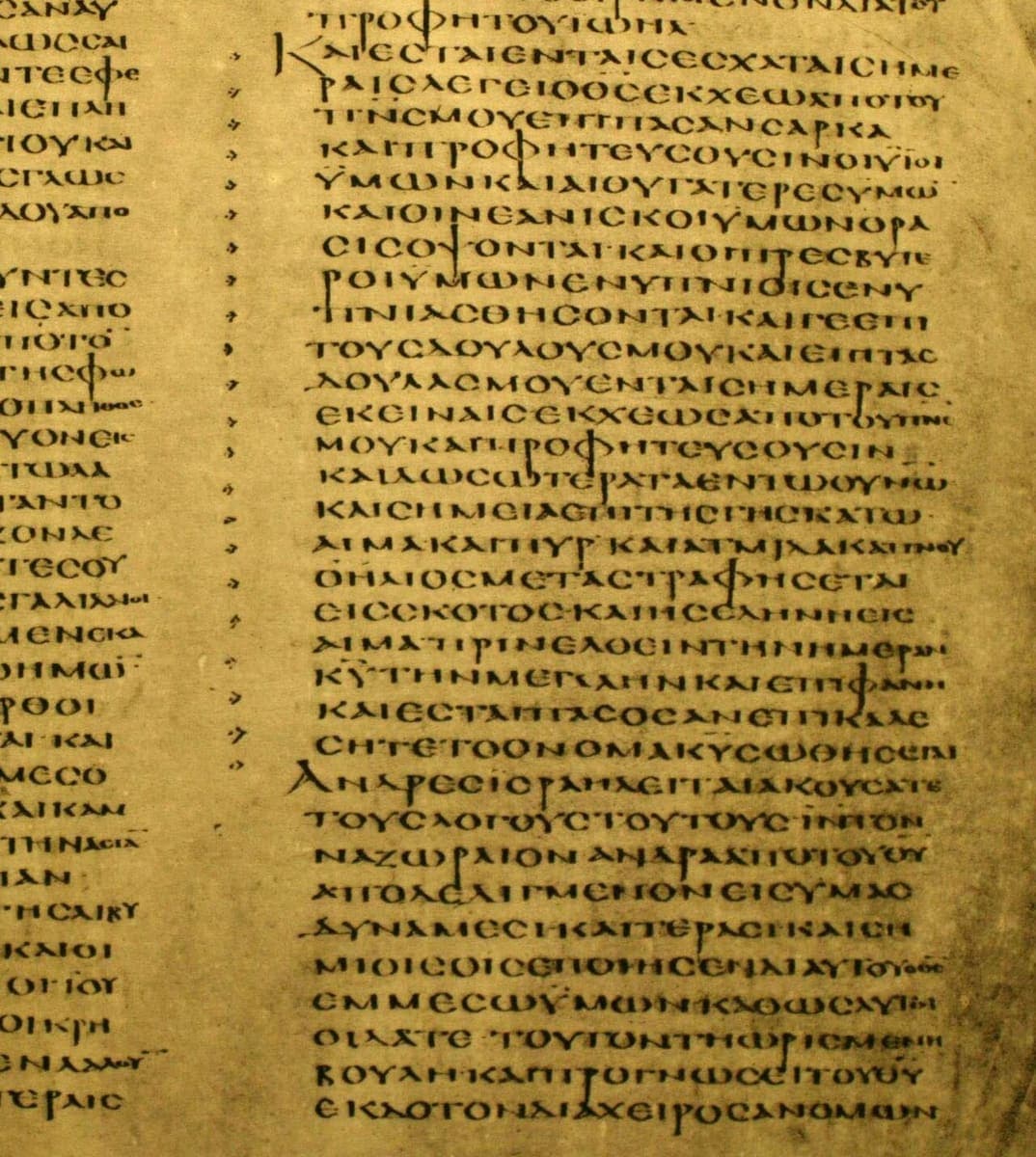 A tall column of fifth-century Greek capitals on yellowed parchment with small arrow-like marks running down the left margin