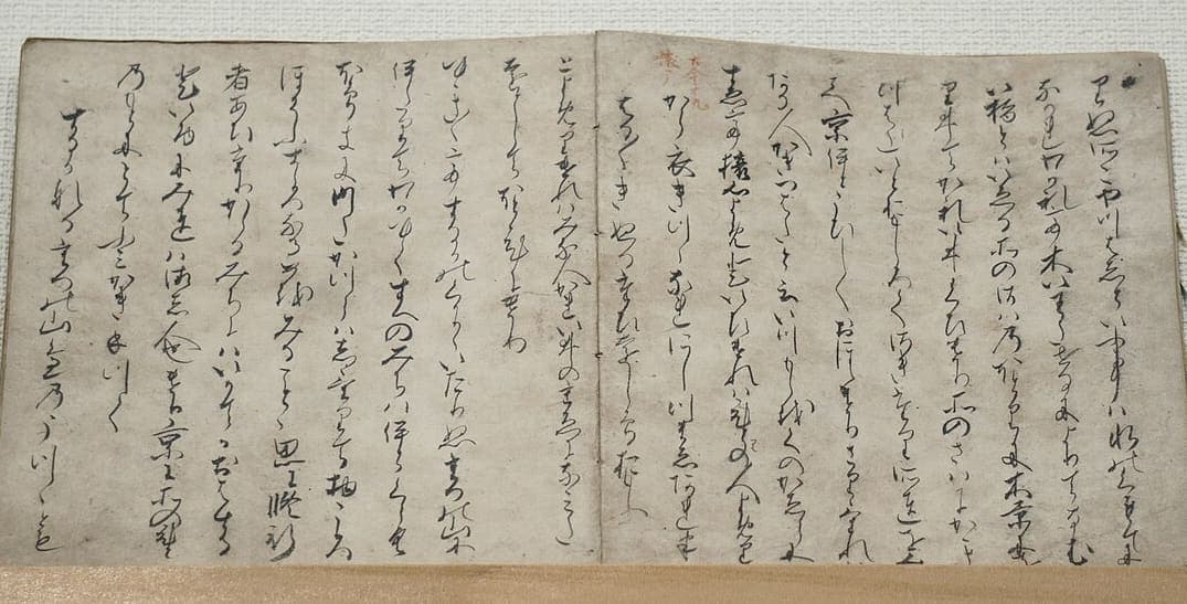 Open double page of a medieval Japanese book, columns of joined cursive kana with a small red section number written in the top margin