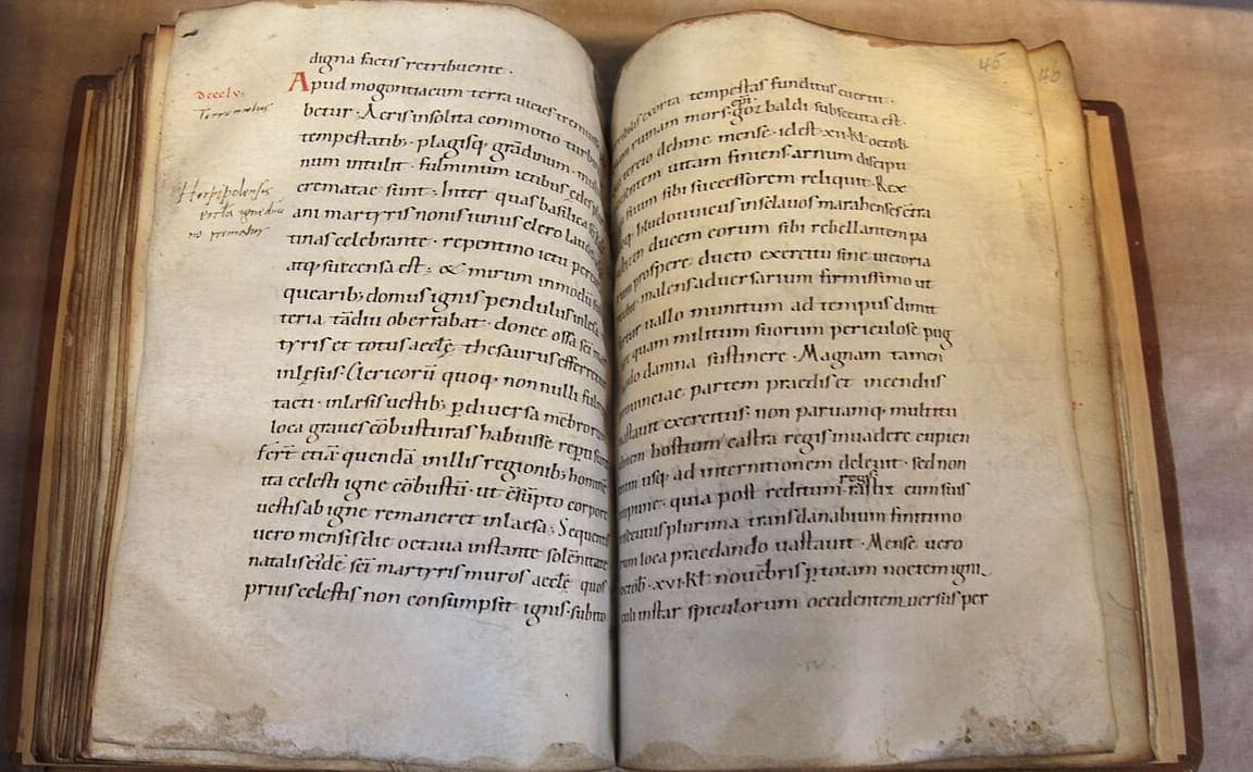 Open parchment book in a museum display case showing two facing pages of dense Latin minuscule annals