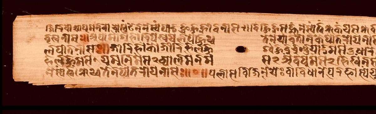 Left portion of a palm leaf with five lines of very small hooked writing, some syllables picked out in red, and a binding hole near the centre